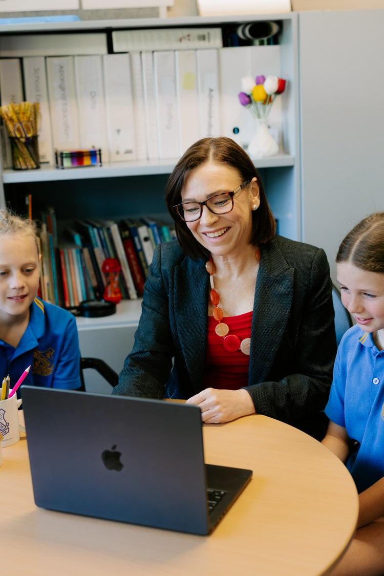 Principal sitting in her office smiling with two students while working on laptop