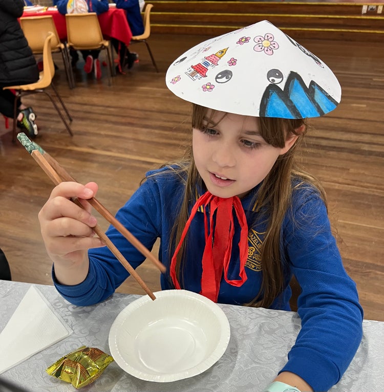 A female student with a colourful hat on and chopsticks for a cultural celebration.