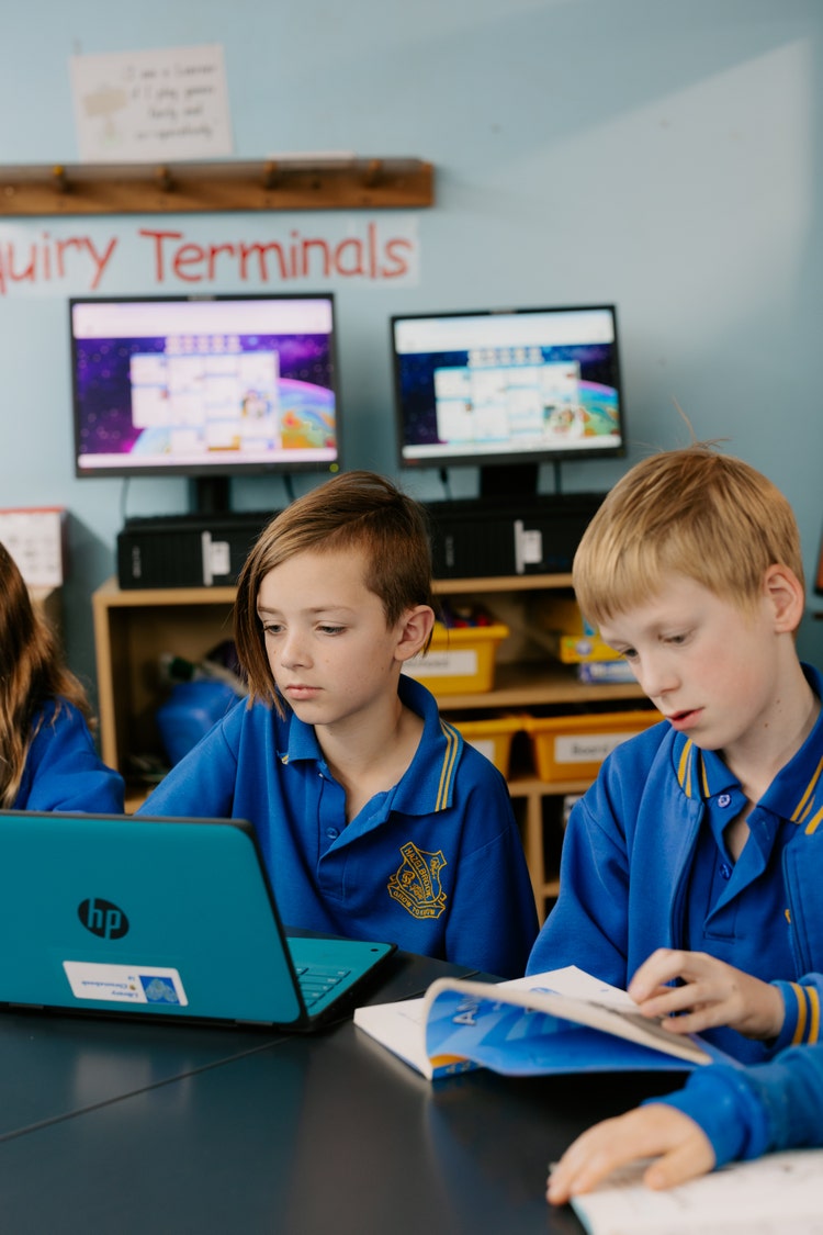 Two male students at a desk. One is looking at a laptop, the other is reading a book
